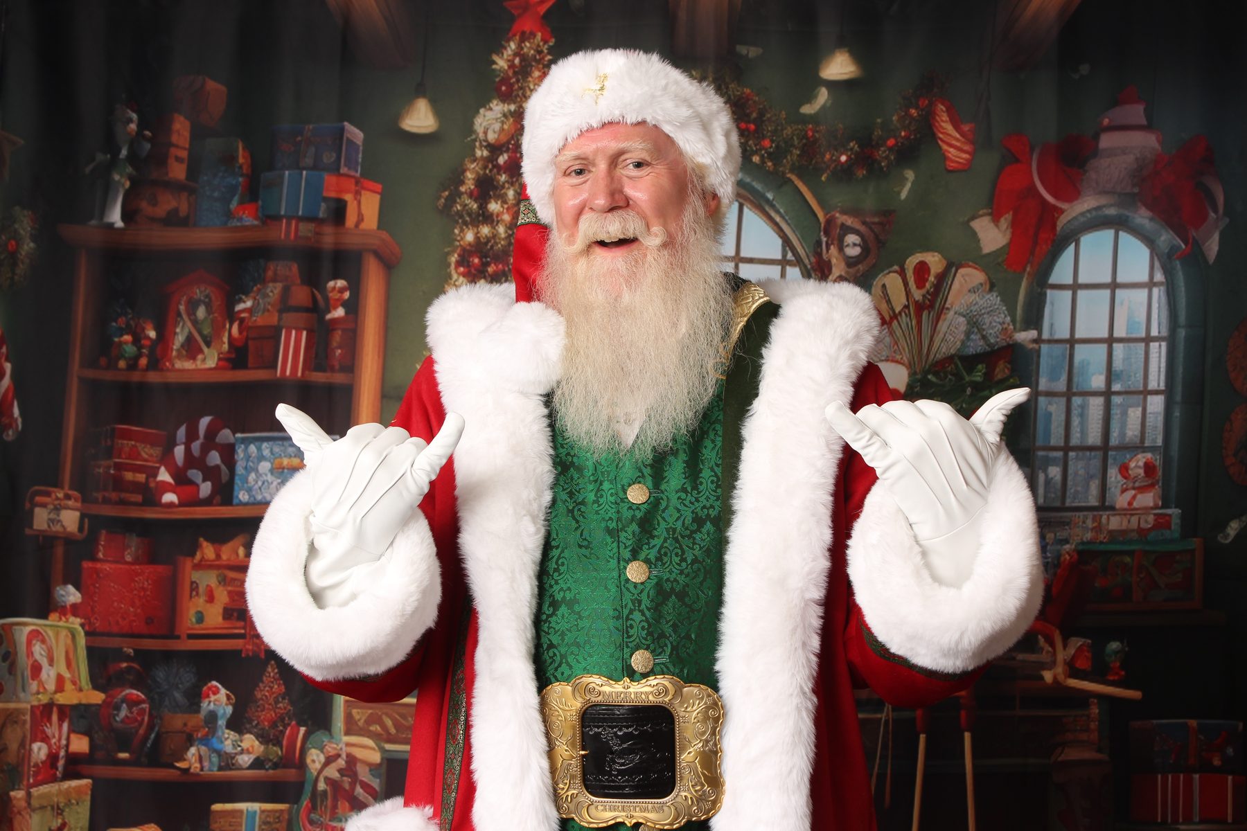 Santa Pat giving a friendly hang-loose gesture with both hands, smiling in a green and red velvet coat