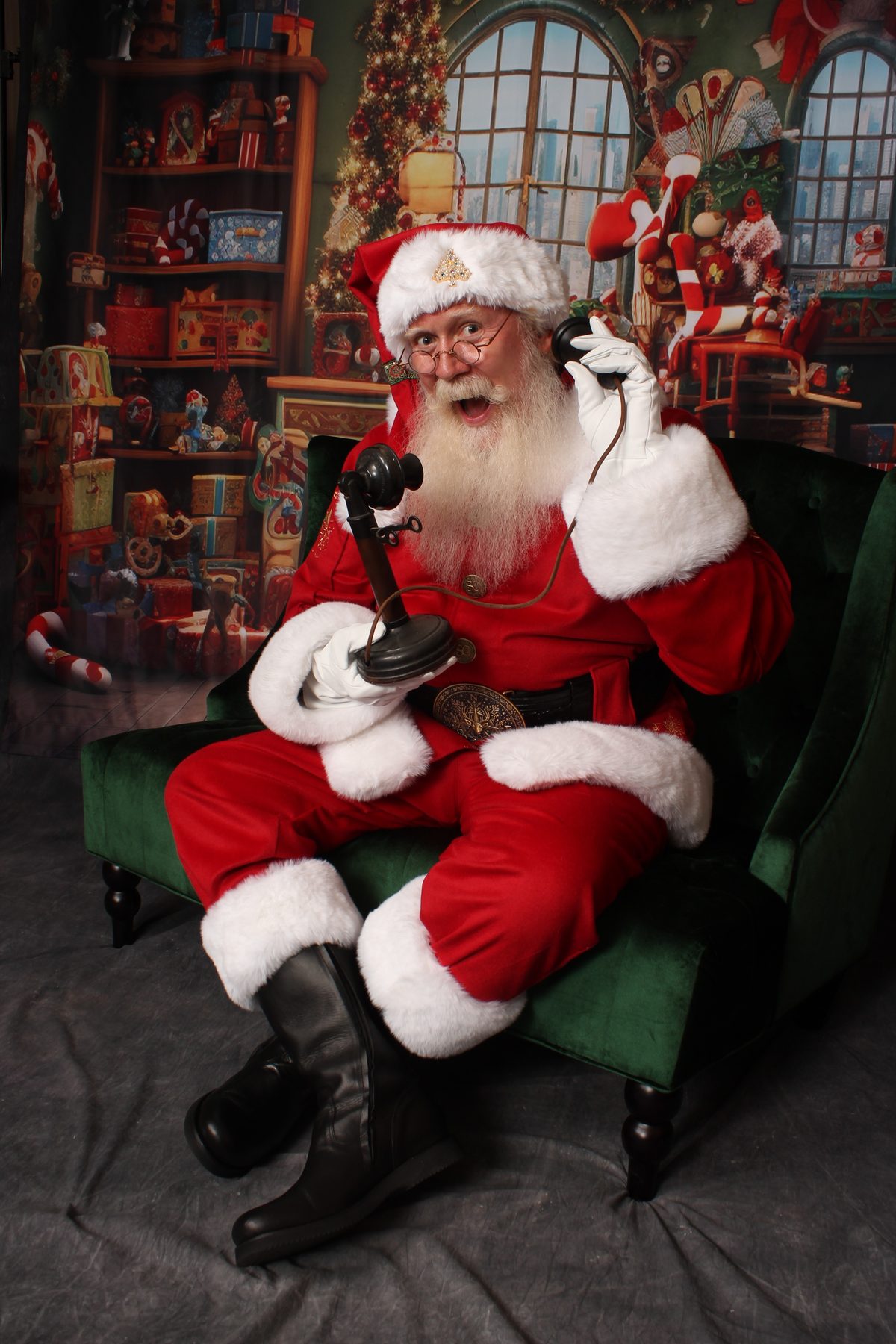 Santa Pat answering an old-fashioned telephone with a delighted expression, seated by a decorated Christmas window
