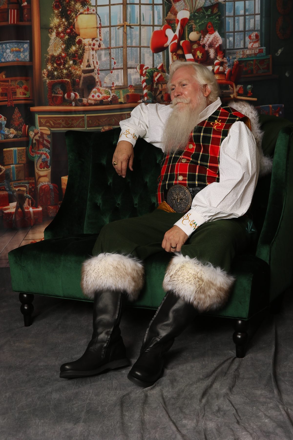 Santa Pat relaxing on a green velvet chaise in his tartan vest and fur-trimmed boots