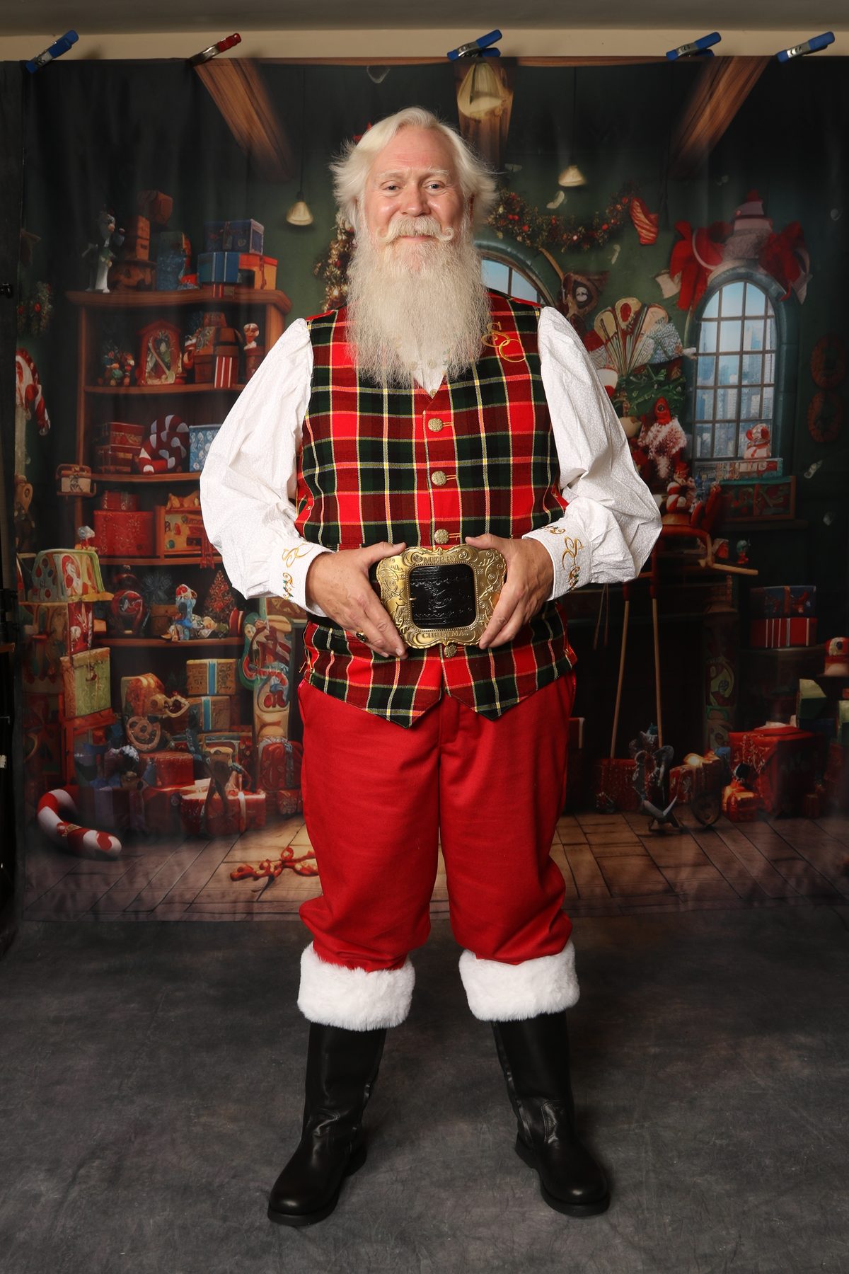 Santa Pat standing in his tartan vest and red trousers in front of a toy-filled backdrop